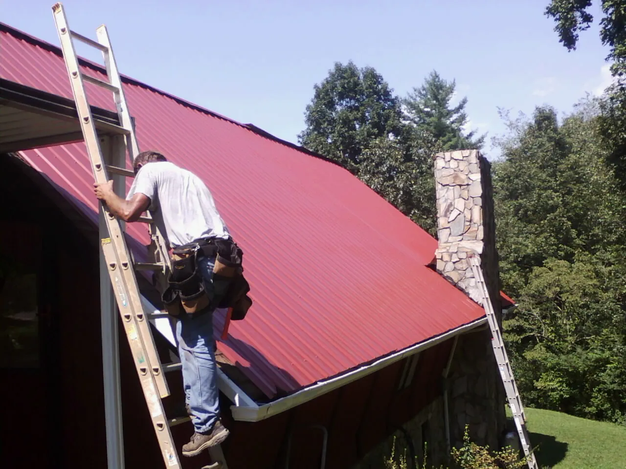 Roofer on a ladder beside a red metal roof and stone chimney — Soffit & Fascia Repair by Roofing Brothers in Elk Horn